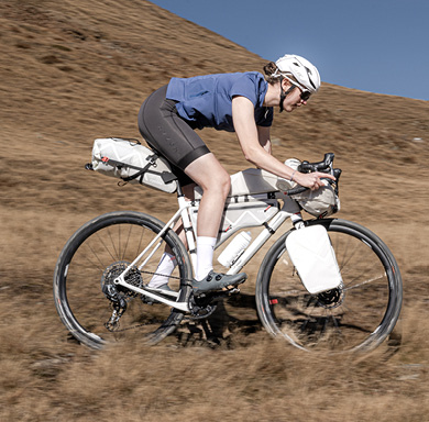 Zu erkennen ist ein Fahrradfahrer auf einem weißen Gravel-Bike in einer bergigen Graslandschaft. Das Gravel-Bike ist mit sehr vielen Fahrradtaschen ausgerüstet.