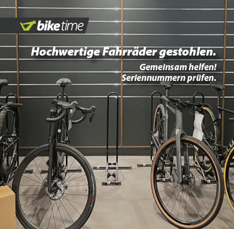 Hochwertige Fahrräder gestohlen. Gemeinsam helfen! Seriennummern prüfen. Zu erkennen sind zwei Rennräder in Bikeständern in einer Verkaufshalle. Zentral in der Mitte ist ein leerer Bikeständer zu sehen.