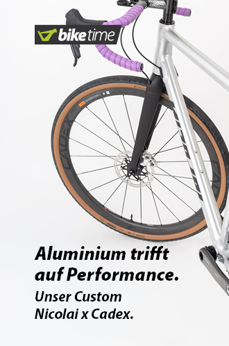 Zu erkennen ist ein Gravel-Bike von hinten mit nach links eingeschlagenem Lenker. Silberner Rahmen, schwarze Laufräder und ein violettes Lenkerband.