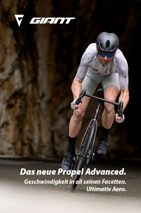Ein Fahrradfahrer fährt auf einem schwarzen Rennrad auf Asphalt. Er kommt direkt auf den Betrachter des Bildes zu. Im Hintergrund ist eine Wand aus Gestein erkennbar.