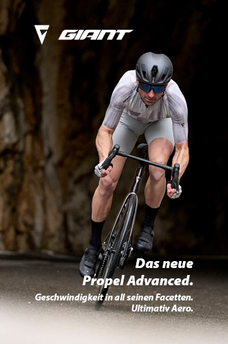 Ein Fahrradfahrer fährt auf einem schwarzen Rennrad auf Asphalt. Er kommt direkt auf den Betrachter des Bildes zu. Im Hintergrund ist eine Wand aus Gestein erkennbar.