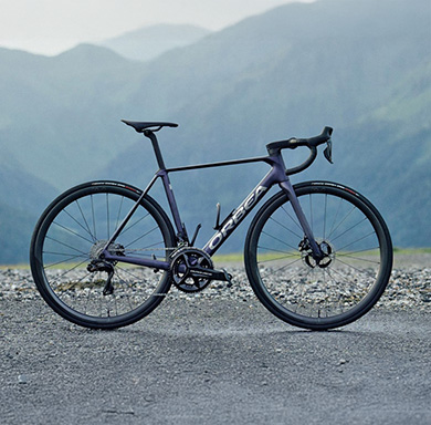 Entdecke jetzt das Orbea Orca! Ein dunkelblaues Rennrad steht auf feinem Schotter vor einer Bergkulisse.