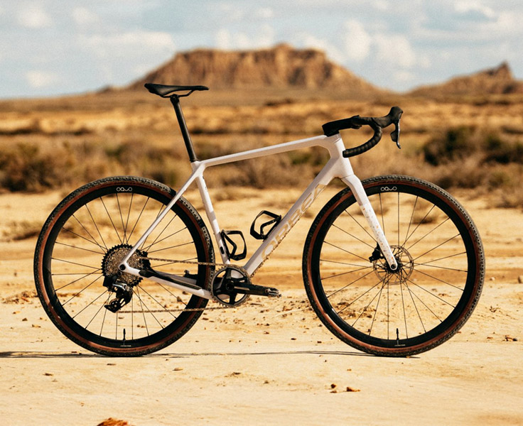 Entdecke jetzt das Orbea Terra! Ein weißes Gravel-Bike steht auf feinem Sand vor einer Wüsten-Kulisse mit Canyons im Hintergrund.