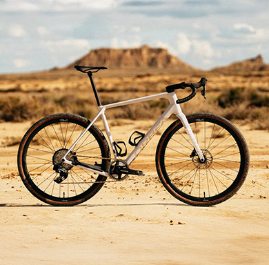Entdecke jetzt das Orbea Terra! Ein weißes Gravel-Bike steht auf feinem Sand vor einer Wüsten-Kulisse mit Canyons im Hintergrund.