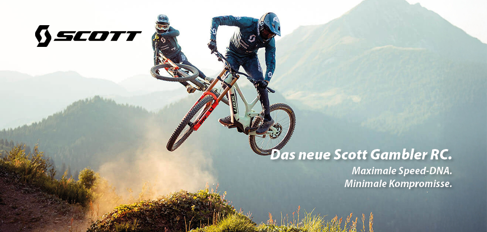 Zu erkennen sind zwei Mountainbiker in hügeligem Gelände die mit ihren Rädern gerade mitten im Sprung sind. Es handelt sich um Mountainbikes der Marke Scott. Das Modell ist das Gambler RC.