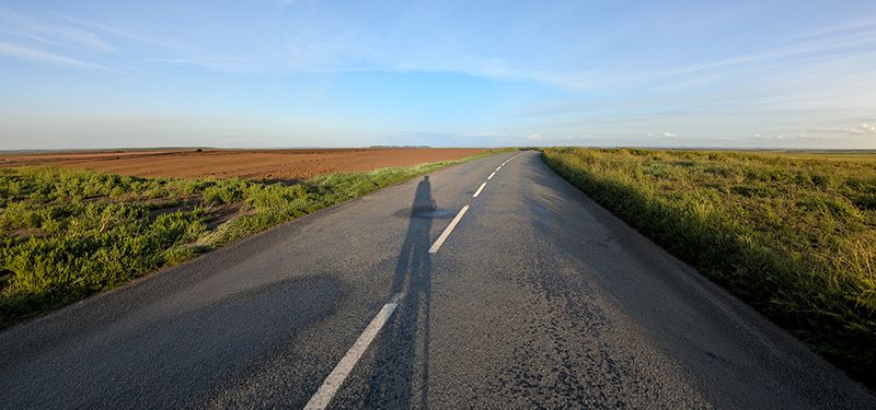 Der Schatten eines Radfahrers senkt sich auf einer flachen geraden Straße, an der landwirtschaftliche Felder angrenzen.