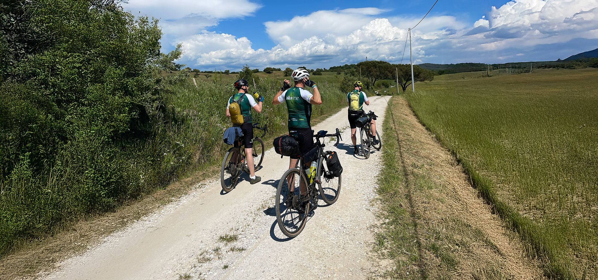 Drei Radfahrer legen auf einer Schotterpiste in einer leicht hügeligen Landschaft eine Pause ein und betrachten die Umgebung.