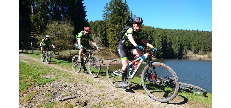Drei Personen auf Mountainbikes fahren durch die Natur. Im Hintergrund ist ein See und Tannenwald zu sehen.