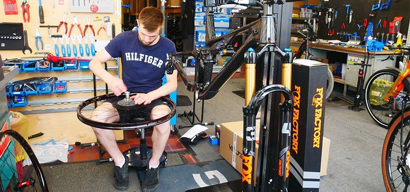 Eine männlich gelesene Person sitzt auf einem Stuhl in einer Fahrradwerkstatt und werkelt an einer Fahrradfelge herum.