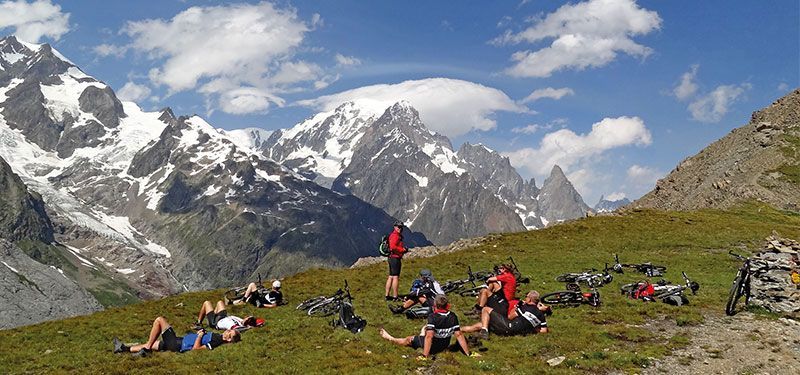 Mehrere Radfahrer entspannen auf einer Bergwiese. Mountainbikes liegen im Rasen. Im Hintergrund sieht man eine große Berkulisse mit Schnee.