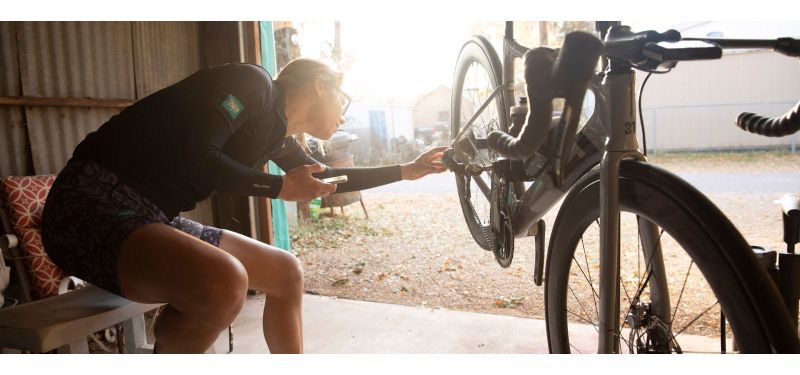 Eine weiblich gelesene Person sitzt auf einer Bank vor einem Fahrrad und kontrolliert etwas am Schaltwerk.