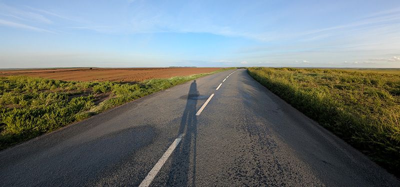 Der Schatten eines Radfahrers senkt sich auf einer flachen geraden Straße, an der landwirtschaftliche Felder angrenzen.