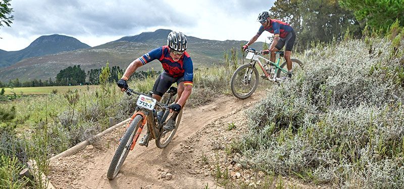 Zwei Mountainbiker auf einer Abfahrt. Im Hintergrund bergige Landschaft. Die Mountainbikes haben vorne Plaketten mit Startnummern von einem Wettbewerb.